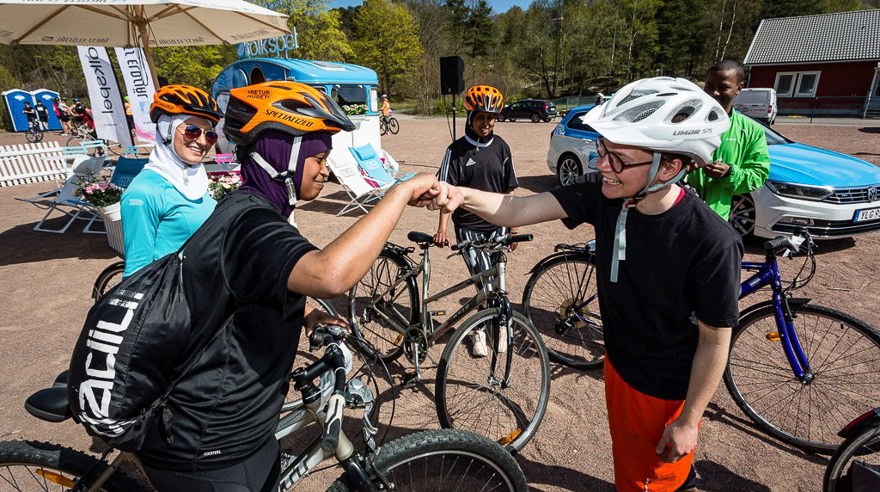 Två kvinnor med cyklar och cykelhjälm hälsar med näven. I bakgrunden andra cyklister och ett skogsområde.
