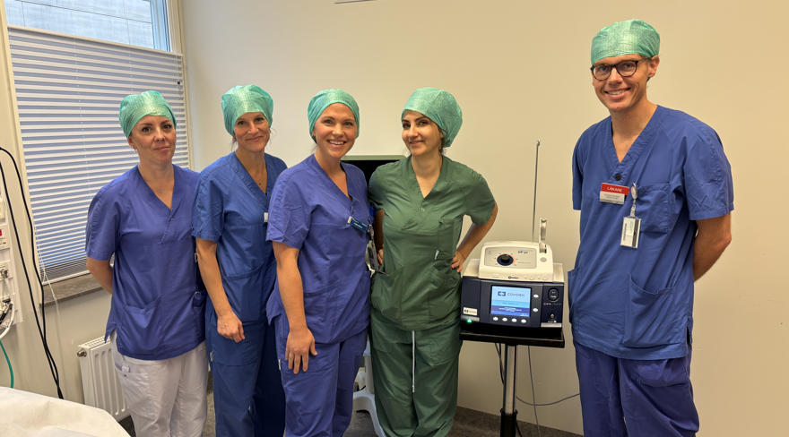 Fem vårdpersonal står samlade i ett operationsrum. Fyra är klädda i blått, en i grönt.