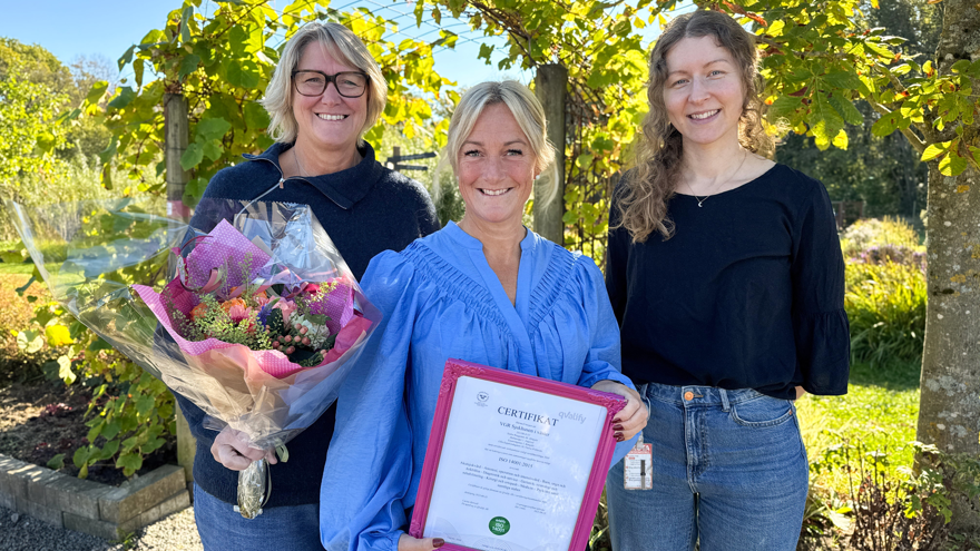Med en bakgrund av färggranna lövträd står tre kvinnliga medarbetare från Sjukhusen i väster. Tillsammans med en blombukett håller personen i mitten i ett certifikat.