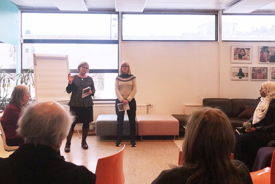Två kvinnor står i ett rum med stora fönster, framför ett blädderblock. I förgrunden sitter personer och lyssnar på presentationen.
