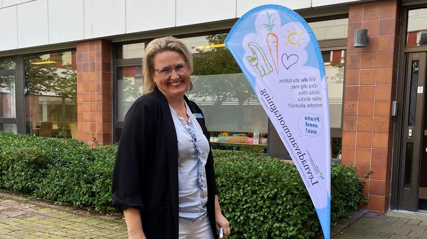 Cecilia ler mot kameran, porten och LeVa-mottagningens beachflagga i bakgrunden.